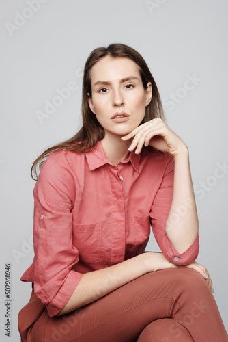 Portrait of dreamy young woman in pink blouse sitting on stool and posing against isolated background