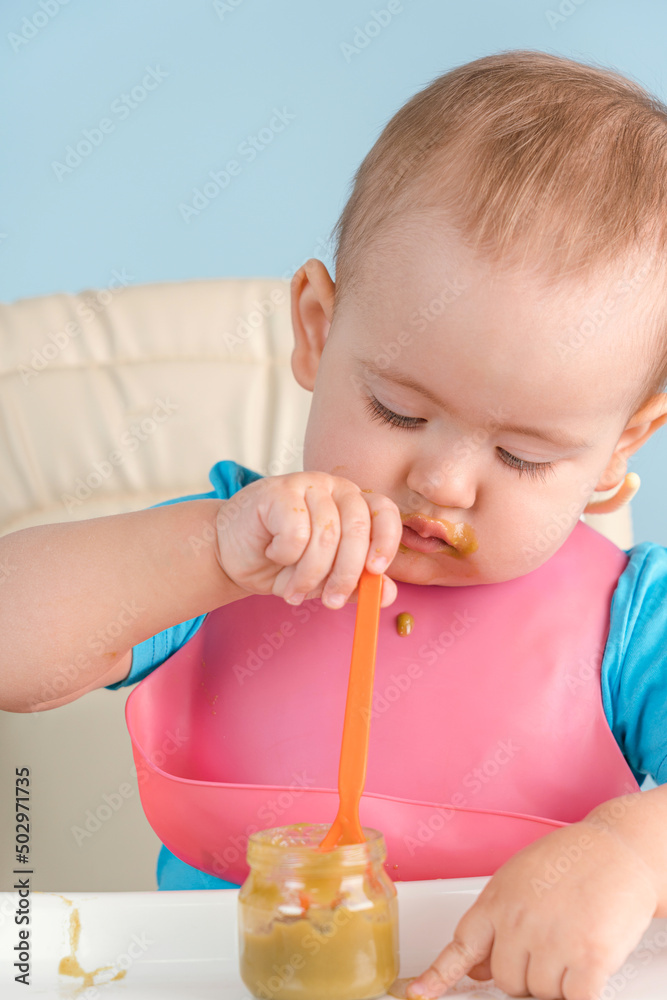 A baby of 12-17 months holding a spoon in his hand eats vegetable mashed broccoli from a glass jar A baby of 12-17 months holding a spoon in his hand eats vegetable mashed broccoli from a glass jar