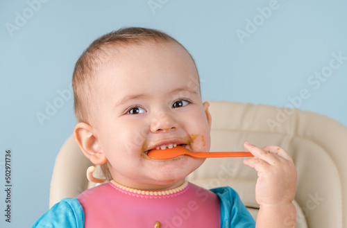 smiling newborn 12-17 months old in a pink bib puts a spoon in his mouth and nibbles it with his teeth