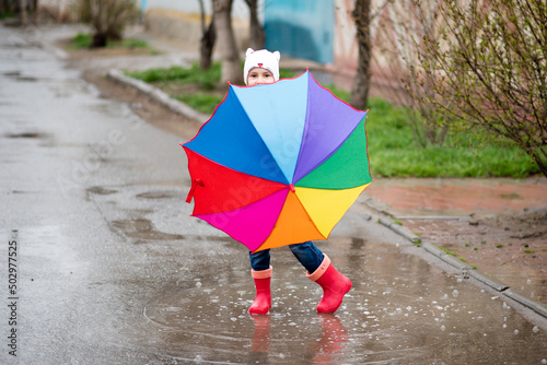 A cute little girl in a red cape  red boots and a white hat jump in puddles and has a fun.The girl has a rainbow umbrella in her hands. Happy childhood. Early spring. Emotions.