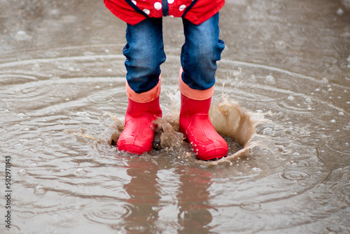 A cute little girl red boots jump in puddles and has a fun. Happy childhood. Early spring. Emotions.