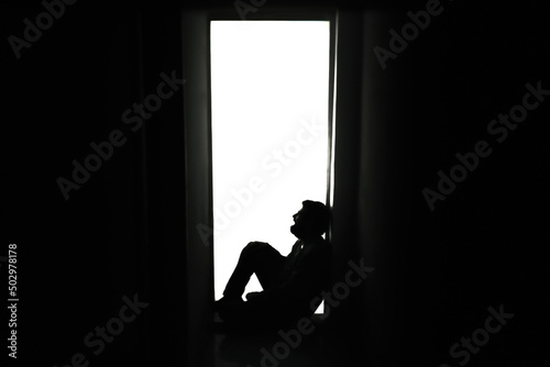 Extreme silhouette shot: a sad, desperate man, crying while sitting alone on the floor near a door in his house, letting his bad feelings come out.
 photo