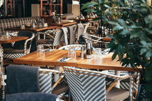 Outdoor empty coffee and restaurant terrace with wooden tables, vintage chairs and warm plaids. Green cafe terrace on the pedestrian street in Europe. A set table is waiting for customers.