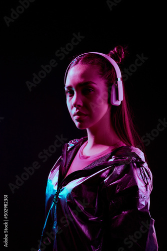 Portrait of serious stylish girl listening to music in wireless headphones while standing in darkness © Mediaphotos