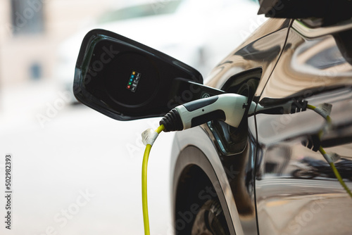 Une voiture électrique est stationnée sur un parking et branchée à une borne de recharge. Le câble de charge est bien visible, reliant le véhicule à la station.