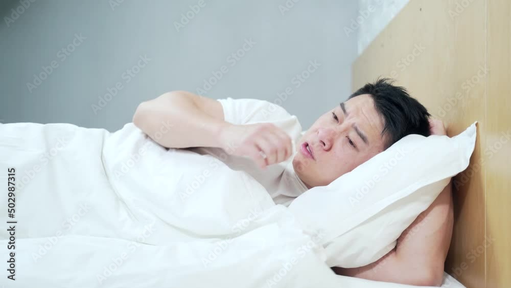 Asian man lying sick in bed with runny nose coughing and flu at home covered with a blanket. Blows her nose into a napkin. Male with fever and headache. Disease. Infection, epidemic, bacillus carrier Asian man lying sick in bed with runny nose coughing and flu at home covered with a blanket. Blows her nose into a napkin. Male with fever and headache. Disease. Infection, epidemic, bacillus carrier