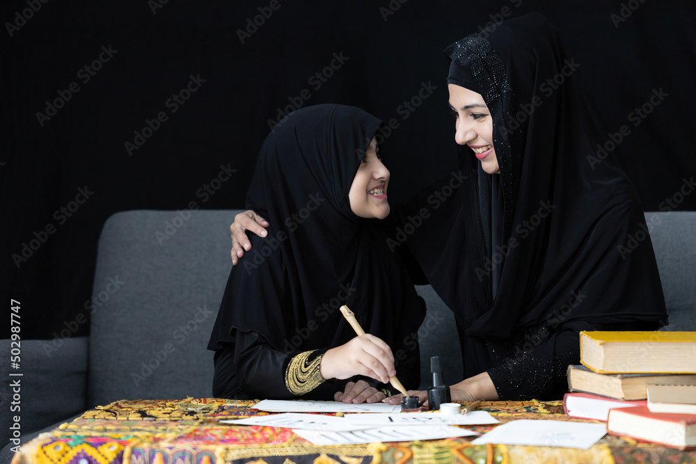 muslim mother with her daughter writing Arabic text with bamboo pens and black ink on paper, Arabic letters mean the name of Muslim god muslim mother with her daughter writing Arabic text with bamboo pens and black ink on paper, Arabic letters mean the name of Muslim god