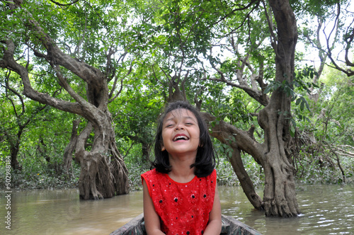 Cute and smiling  little girl wearing red dress sitting on boat with closed eyes and enjoying vacation at  Ratargul Swamp Forest trees background.  Travel vacation lifestyle concept. photo