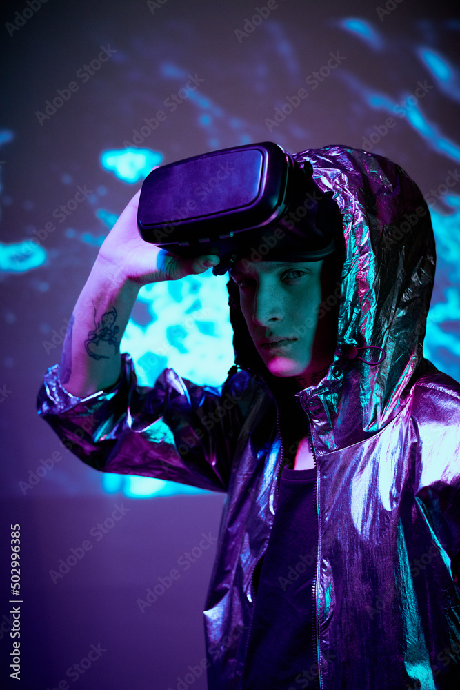 Portrait of serious young male VR gamer in hooded jacket taking off virtual reality goggles