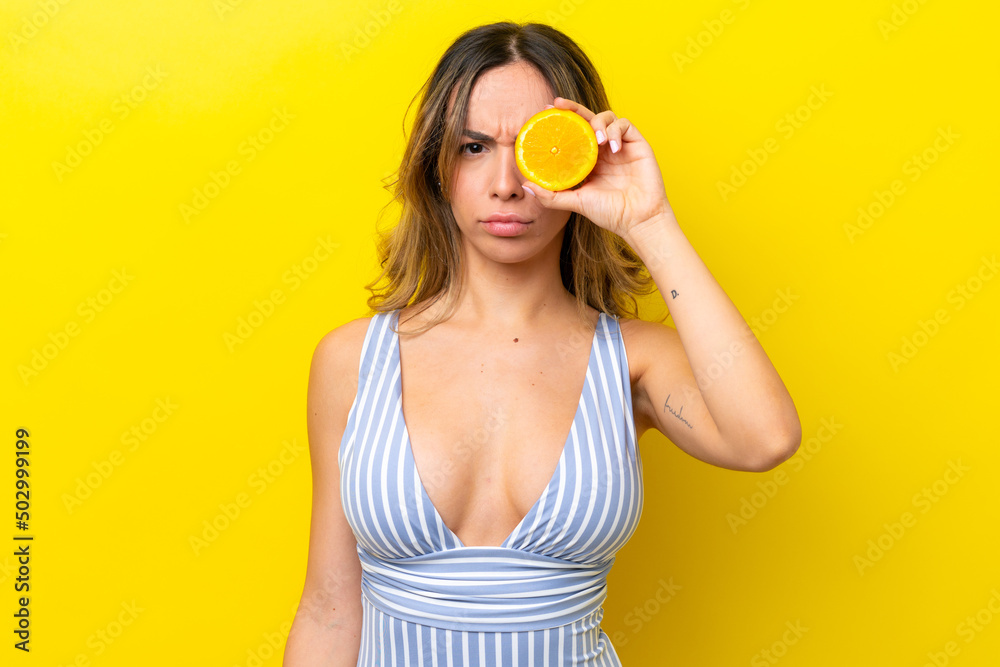 Young caucasian woman isolated on yellow background in swimsuit and holding an orange Young caucasian woman isolated on yellow background in swimsuit and holding an orange