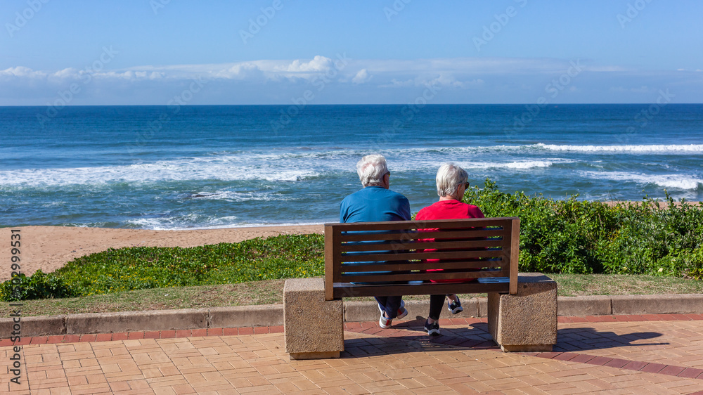 Mature Man Woman Couple Unrecognizable Sitting On Bench Promenade Pathway looking View Of  Beach Ocean Landscape
