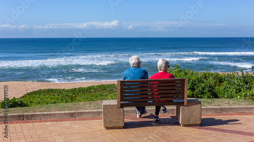 Mature Man Woman Couple Unrecognizable Sitting On Bench Promenade Pathway looking View Of Beach Ocean Landscape