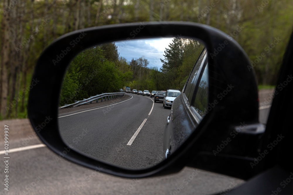 The mirror of a passenger car, which reflects a traffic jam on a country road in summer.