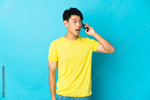 Young Chinese man isolated on blue background keeping a conversation with the mobile phone