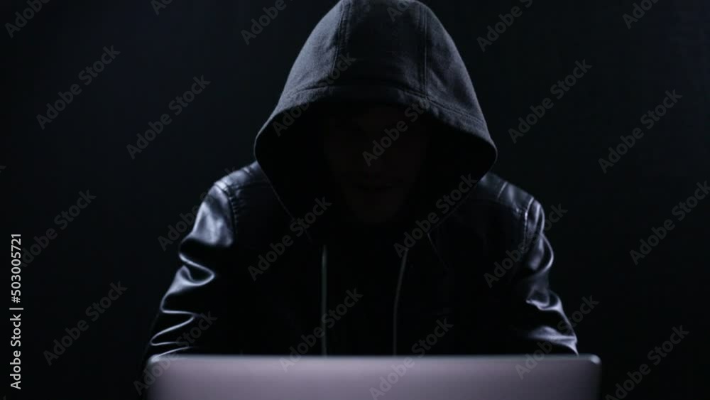 Hooded figure typing on a laptop in the dark Hooded figure typing on a laptop in the dark