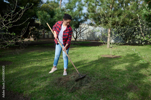 Full length of female gardener raking grass clippings into a compost heap in her garden of a countryside house backyard