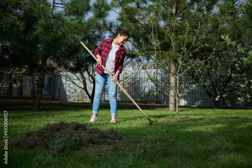 Young woman gardener cleans the local area, rakes the cut grass into a pile for further preparation of compost for fertilizing the black soil