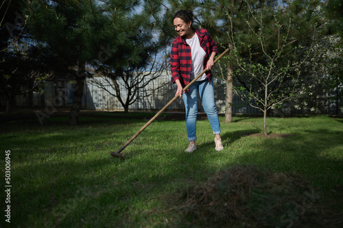 Full length a charming Hispanic woman gardener cleaning the local area and raking grass against blossoming fruit tree background on a sunny spring day