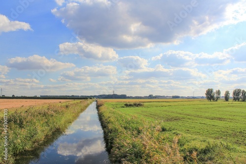 a dutch rural landscape with a broad ditch between green fields and a blue sky full of white clouds in springtime