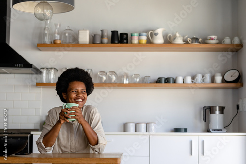 Smiling mid adult african american woman looking away while having coffee in kitchen at home photo