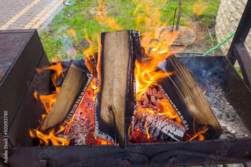 Birch firewood burns with red flames in a black metal grill on a sunny summer day