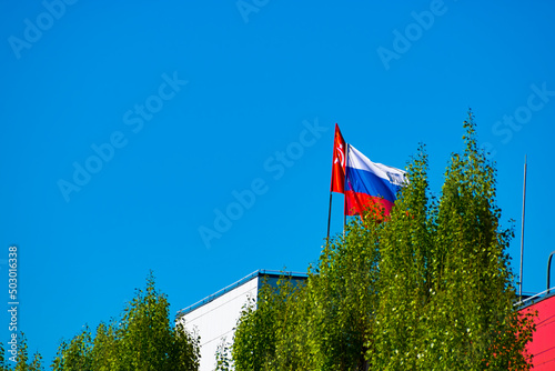 he Russian flag next to the victory banner on the roof of the building behind a tall tree.
