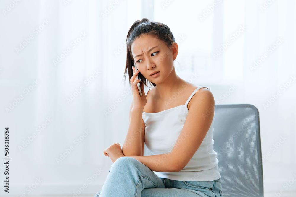 Wary irritated angry tanned lovely young Asian woman listen bad news at call using phone at home interior living room. Distance communication Social media concept. Cool offer Banner Wide angle Wary irritated angry tanned lovely young Asian woman listen bad news at call using phone at home interior living room. Distance communication Social media concept. Cool offer Banner Wide angle