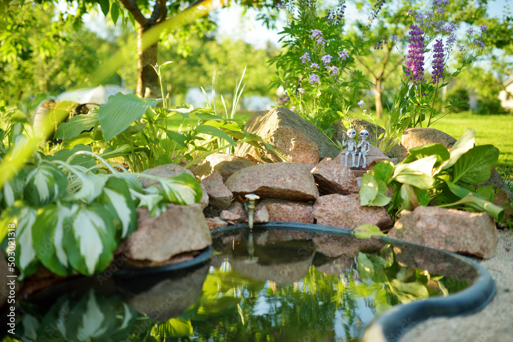 Small artificial pond on sunny summer day in the garden. Beautifully designed garden pond surrounded with plants and flowers. Small artificial pond on sunny summer day in the garden. Beautifully designed garden pond surrounded with plants and flowers.