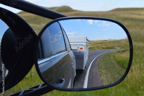 Side rearview mirror reflects side of black vehicle and the pop-up camper being towed, blue skies, long curving road, and grasslands in South Dakota. © Kathryn