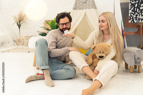 Inner shot of a caucasian excited couple in their 20s sitting on the floor in a nursery with their legs crossed. Woman holding a small baby boot and a giant teddy bear enjoying future motherhood. High