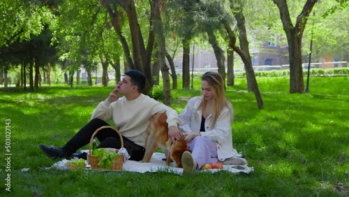 A young man smokes a cigarette on a picnic with his girlfriend, the girl waves her hand from the smoke. A young couple is sitting in the park on a picnic and stroking their dog on the head. Picnic in