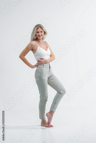 A beautiful blonde with a sporty figure in light jeans and a white T-shirt poses in the studio on a white background