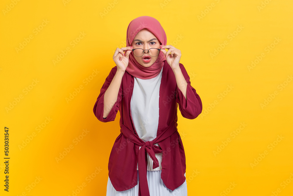 Fototapeta premium Amazed beautiful Asian woman in casual shirt looking at camera taking off eyeglasses, shocked by news or special offer isolated over yellow background