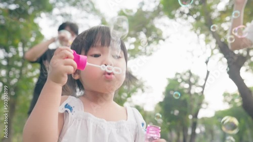 Asian girl blowing soap bubbles having fun playing with parents outside, Asian child innocent and curiosity, little preschool kid, family enjoy happy time in the park under trees shades