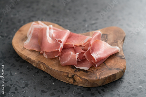 Thin italian prosciutto slices on olive wood board on conctere background