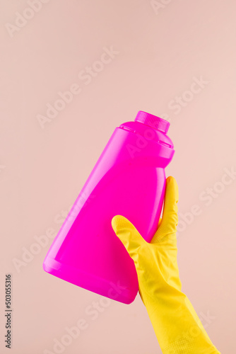 Hand in protective glove holds bright pink bottle of detergent on beige closeup