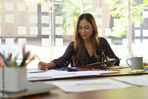 Thai Asian female working on her design developer career while writing something on paper with focused face in the modern office interior. Creative digital development concept.