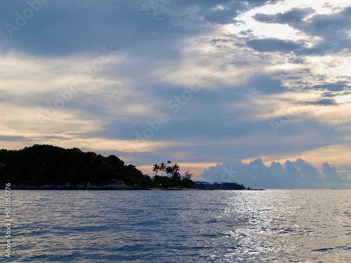 Sunset over tropical island. Picturesque illustration. Photo of sundown. View from the sea. Silhouette of island and several palm trees against the sunset sky. Amazing clouds in the skies. Sun glow
