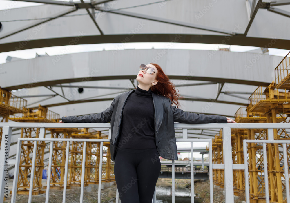Red-haired woman in sunglasses and a black leather jacket against the background of an urban industrial landscape
