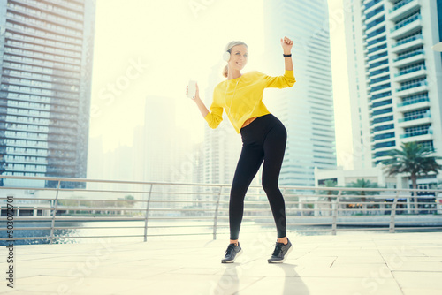 Sport and music. Happy young woman with earphones and smartphone dancing while exercising on city embankment. © luengo_ua