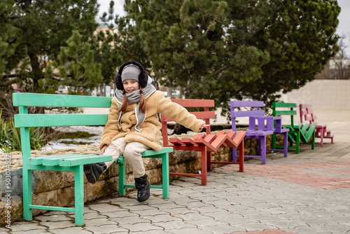 a cute six-year-old girl in a beige fur coat made of eco-friendly faux fur is sitting on a bench