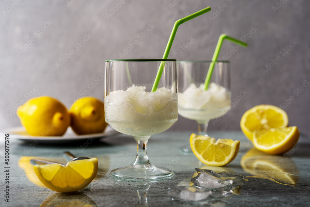 Lemon sorbet in two glasses with lemons, ice cubes and drinking straws on grey background Lemon sorbet in two glasses with lemons, ice cubes and drinking straws on grey background