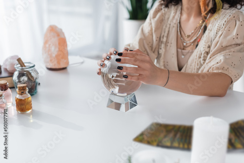 Cropped view of fortune teller touching orb near blurred tarot cards and witchcraft supplies on table.