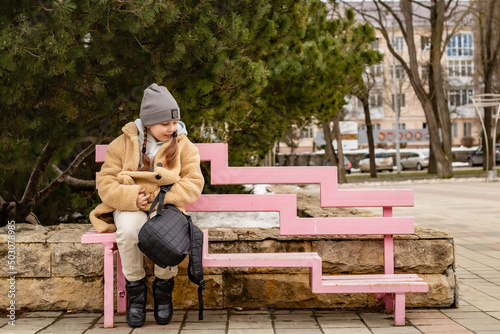 a cute six-year-old girl in a beige fur coat made of eco-friendly faux fur is sitting on a bench