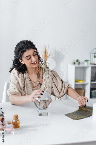 Smiling gypsy medium touching magic orb and tarot cards on table.