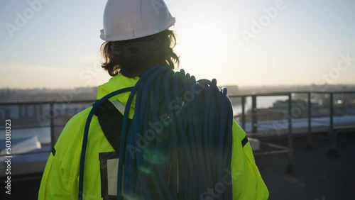 Professional climber in working outfit and helmet. Industrial climber with skein of blue rope walks to rooftop edge with city view at sunset closeup photo