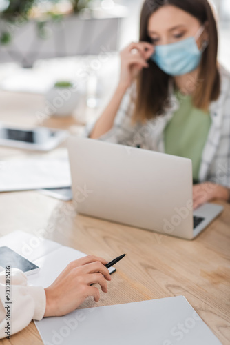 Businesswoman holding pen near notebook  cellphone and blurred colleague in office.