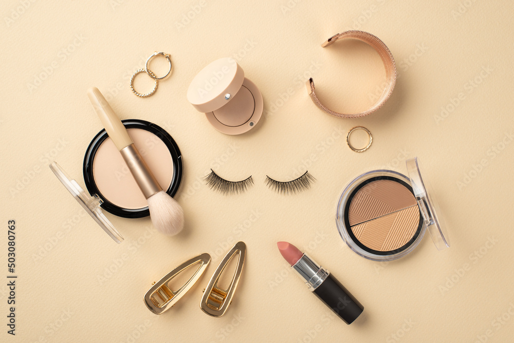 Make up concept. Top view photo of circle composition false eyelashes lipstick compact powder brush contouring palette eyeshadow gold wristlet rings and barrettes on pastel beige background Make up concept. Top view photo of circle composition false eyelashes lipstick compact powder brush contouring palette eyeshadow gold wristlet rings and barrettes on pastel beige background