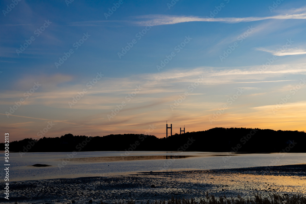 Sunset with reflections in water and silhouette of treeline. Bridge pylons in distance.