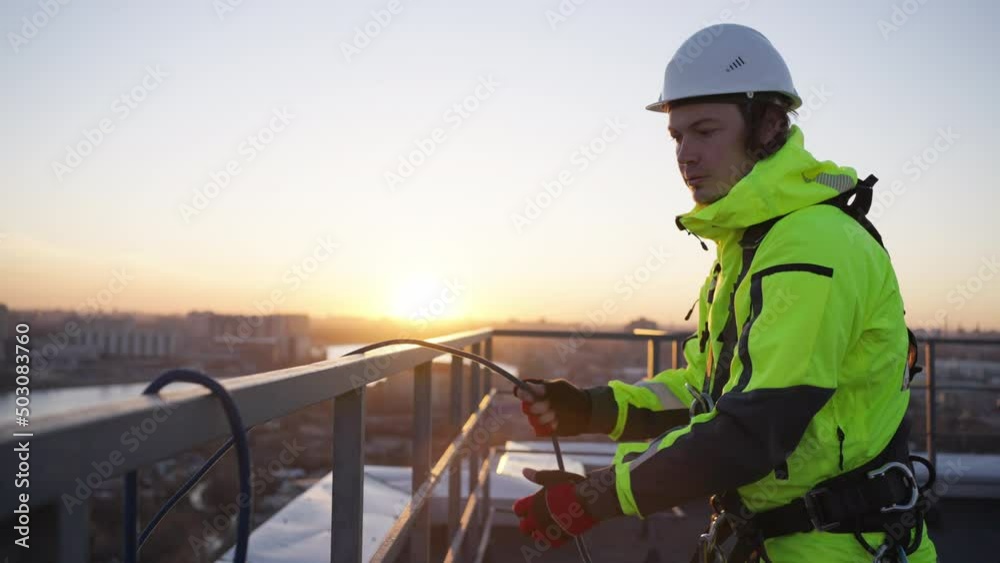 Industrial climber removes safety equipment from roof fence at sunset. Man in green overall finishes work on rooftop of skyscraper in evening Industrial climber removes safety equipment from roof fence at sunset. Man in green overall finishes work on rooftop of skyscraper in evening